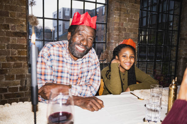 Black family members sitting together at dining table, wearing paper crowns, and grinning at camera. On-camera flash, retro-style photographic effect.