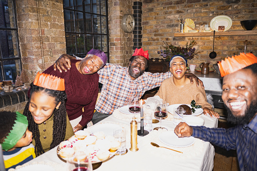 Festive adults and children wearing paper crowns, gathered around dining table, grinning at camera. On-camera flash, retro-style photographic effect.