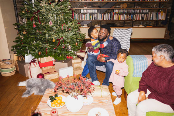 Young son giving father Christmas present, hug, and kiss Mid adult Black man, children, and their grandmother together in loft apartment and celebrating the holiday. On-camera flash, retro-style photographic effect. pics of the black granny stock pictures, royalty-free photos & images