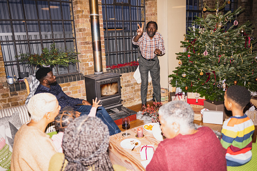 Senior Black man standing in front of adults and children seated in cozy loft, all watching him give clues. On-camera flash, retro-style photographic effect.