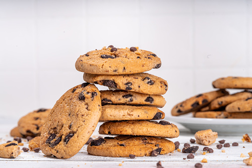 Top view of a wooden plate filled with chocolate chips cookies placed at the center of the frame. Ingredients for preparing and baking cookies are all around the plate. Some baked cookies are on a cooling rack. Ingredients includes are flour, eggs, chocolate chips, brown sugar and butter. Some kitchen utensils complete the composition. Predominant colors are brown and blue. Low key DSRL studio photo taken with Canon EOS 5D Mk II and Canon EF 100mm f/2.8L Macro IS USM