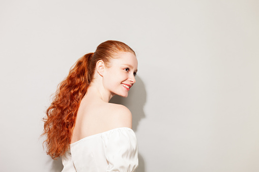 Studio portrait of a young attractive cheerful red-haired woman in a white blouse against a gray background