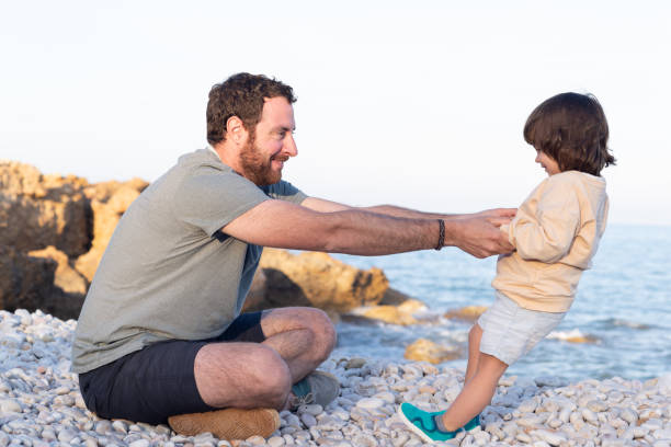 istock Father holding hands to her little daughter while she is balancing backwards on a pebble beach at sunset. 1486054102