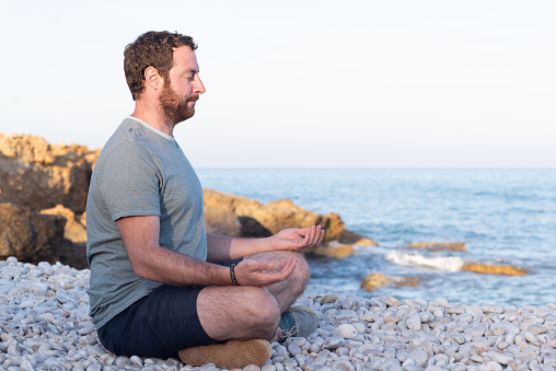 Profile view of a smiling man with eyed closed sitting in Sukhasana yoga pose on a pebble beach at sunset.