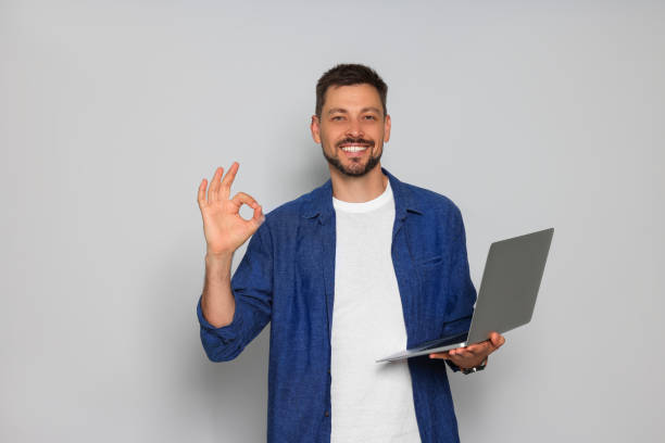 istock Smiling man with laptop showing okay gesture on light grey background 1486055497