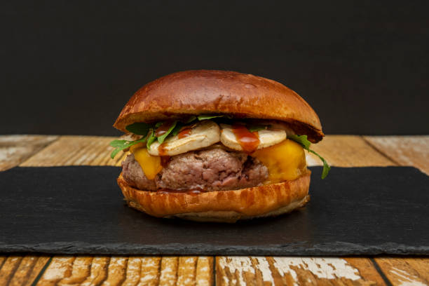 istock beef burger with arugula, mushrooms, barbecue sauce and cheddar cheese on a black slate plate and unpainted wooden table 1486058769