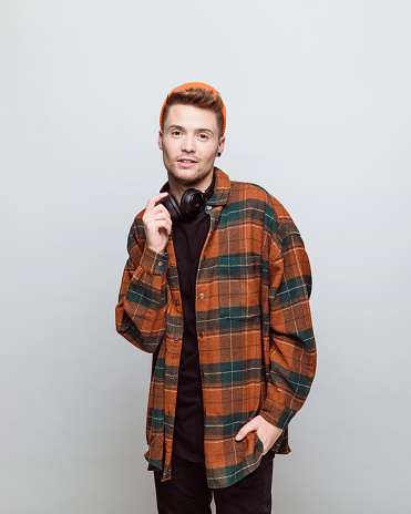 Portrait of young man wearing orange beanie and checkered shirt smiling at camera. Studio shot, grey background.