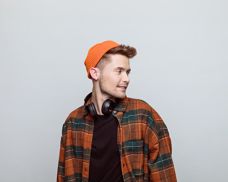 Portrait of young man wearing orange beanie and checkered shirt looking away. Studio shot, grey background.