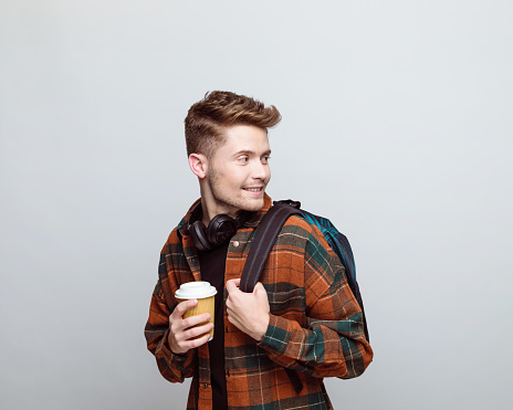 Portrait of young man wearing checkered shirt and backpack, looking away and holding take away cup of coffee. Studio shot, grey background.