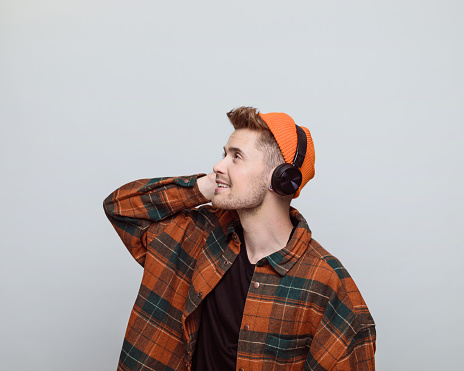 Portrait of young man wearing orange beanie, headphone and checkered shirt listening to the movie. Studio shot, grey background.
