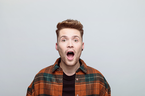 Portrait of excited young man wearing orange checkered shirt looking at camera with mouth open. Studio shot, grey background.