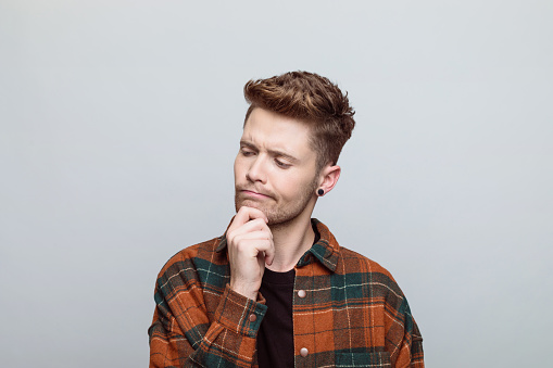 Portrait of thoughtful young man wearing orange checkered shirt contemplating with hand on chin. Studio shot, grey background.