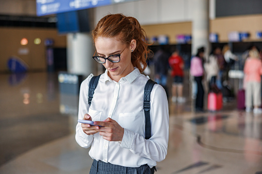 Waist-up front shot with blurred background of a pretty ginger mid-adult Caucasian businesswoman texting on her phone while waiting to be called to board the plane for her first business trip at the airport during a sunny day.