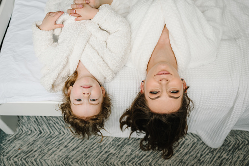 Young mother with cute little daughter in bathrobe lying on bed, lovely look at camera. Healthcare and family concept. Happy mom and girl kid smiling top view. Cheerful girls having beauty day at home