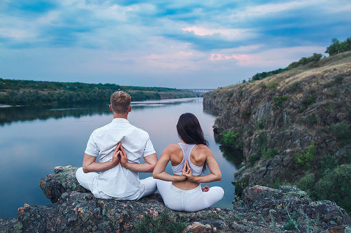 Rear view of a guy and a girl doing yoga in the Pashchima Namaskarasana pose on the river bank