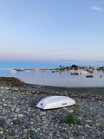 Rye Harbor, United States – August 28, 2022: A flipped boat on the rocky shore against the background of the coast with vessels. Rye Harbor Beach.