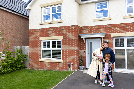 Wide shot of a mid-adult couple standing with their two children in front of their family home. They are all laughing together wearing casual clothing and looking into the camera.
