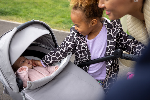 Over-the-shoulder shot of a newborn baby girl in a pram. Her older sister is reaching into the pram checking on her.