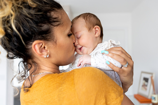 Waist-up shot of a mid-adult woman holding her newborn daughter. The mother is wearing casual clothing and kissing her daughter both closing their eyes.