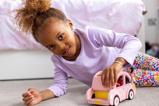A three-quarter length of a young female child playing with a toy car in her room. She is looking into the camera.