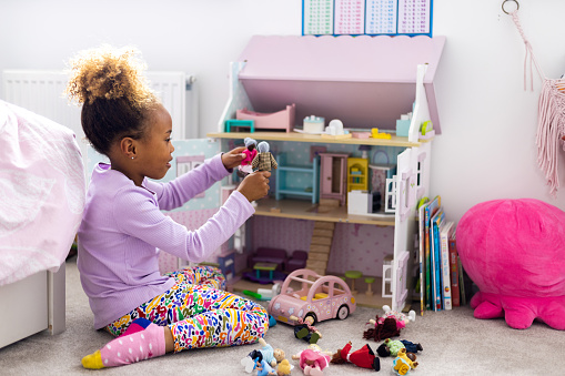 A side view full shot of a young female child playing with a doll's house in her room. She is playing with dolls in her hands kneeling on the floor.