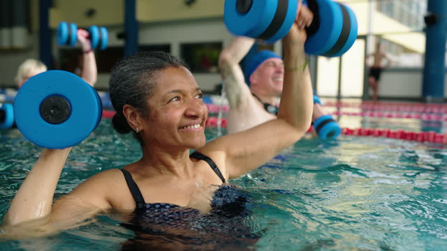 Active senior woman with aqua dumbbells exercising at water aerobic session in pool
