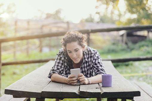 Lying on a wooden plank in the countryside or in the mountains, a woman who loves adventure tourism enjoys the freedom of nature but stays in touch with others with her smartphone.