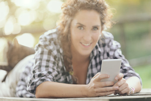 Lying on a wooden plank in the countryside or in the mountains, a woman who loves adventure tourism enjoys the freedom of nature but stays in touch with others with her smartphone.