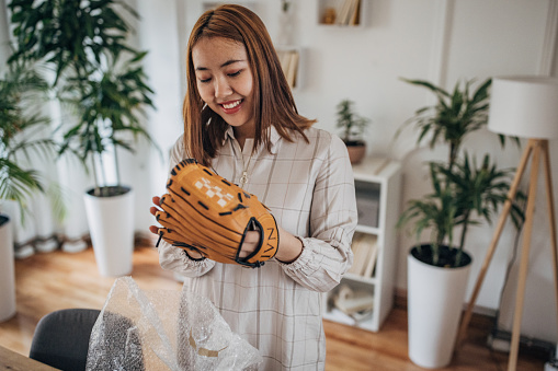 Young Asian woman opening a package at home, she ordered a baseball glove.