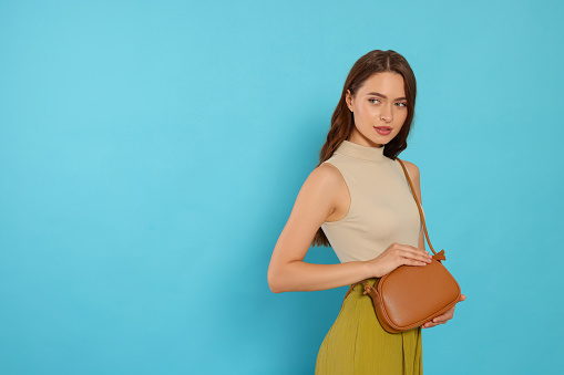 Beautiful young woman in fashionable outfit with stylish bag on light blue background, space for text