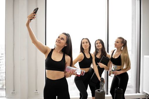 Attractive healthy women with sports bottles taking selfie on cell phone while resting after yoga class in fitness centre. Cheerful friends in modern activewear enjoying healthy lifestyle together.