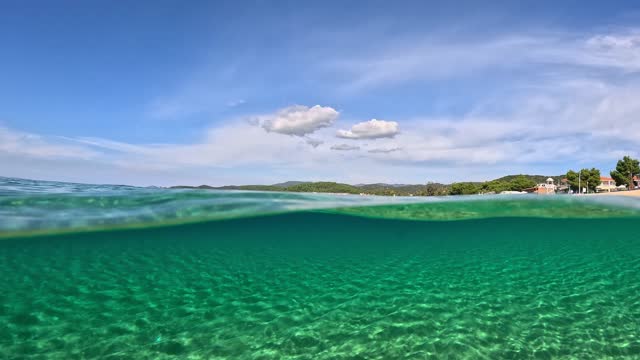 Crystal clear sea water in Sitonia, Halkidiki, Greece. Half underwater slow-motion view of sandy beach bottom, blue sky and clouds. Shot from border of air and water with dome