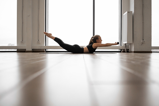 Active caucasian woman lying downward facing on yoga mat and bending back with dumbbells in hands. Strong young blonde in sport clothes having intense workout in airy room.
