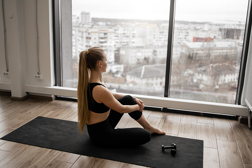 Healthy fit young woman in sport clothes enjoying amazing city view from panoramic window while resting after workout at spacious room. Concept of people, body care and recreation.