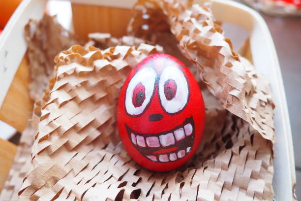 istock Easter egg painted red, white and black with a cheerful face. Boiled chicken dyed egg with red shell. Fun background. Big eyes, white teeth on the face. Easter basket with cut craft paper. 1486133053