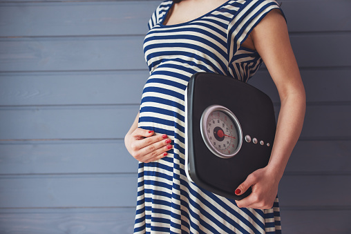 Cropped image of beautiful pregnant woman holding weigh scales and keeping one hand on a belly, standing against gray wall