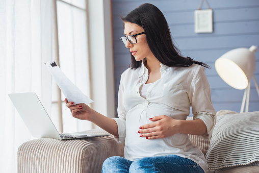 Beautiful pregnant business woman in eyeglasses is examining documents and using a laptop while working at home