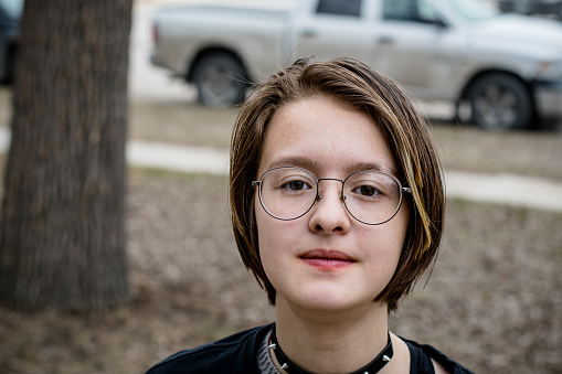 This portrait captures the essence of a 15-year-old gender non-conforming individual who boldly expresses themselves through their punk attire. With short hair and a blond highlight on the front, they stand out with their large glasses and shy smile. This image celebrates individuality and self-expression, breaking free from traditional gender norms. It is a powerful representation of the LGBTQ+ community and their struggles for acceptance and recognition.