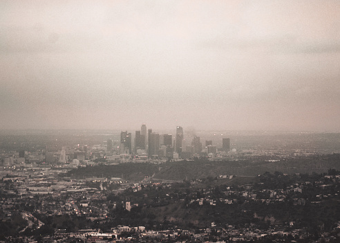 Photo taken from the window of a Cessna 172. Downtown Los Angeles captured in a vintage film aesthetic. The towering skyscrapers and fancy surrounding neighborhoods make up the famous City of Angels cityscape view. Building lights twinkle in the blue hour lighting. Haze shrouds the surrounding environment on a gloomy day.