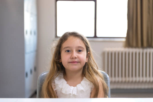 Photo of a little tired girl in a pink blouse bowing her head while sitting at a wooden table behind a large glass window in her room. Difficult childhood concept