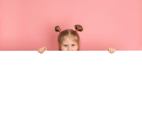 Funny girl hiding behind blank white advertising billboard with copy space and peeking out pink wall.