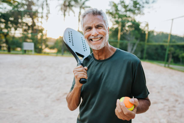 älterer mann, der strandtennis spielt, schaut in die kamera - tennis active seniors healthy lifestyle senior men stock-fotos und bilder