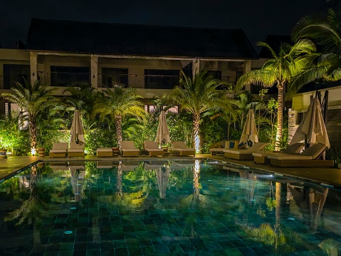 An outdoor swimming pool illuminated with bright lights surrounded by lush palm trees, providing a tranquil setting