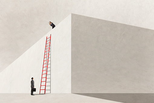 A businessman looks at a tall ladder leaning against a wall as a businesswoman who has already climbed the ladder looks back down at him.