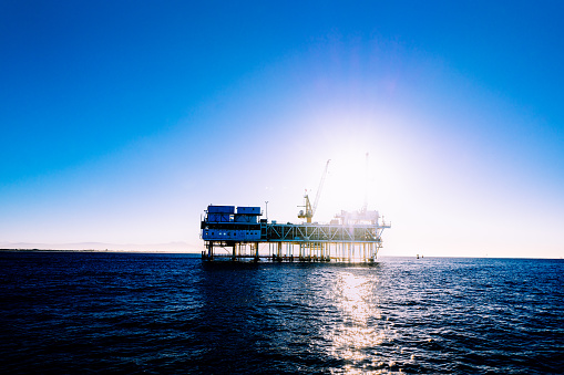As the sun rises over the Pacific, the silhouette of a massive offshore oil rig stands in stark contrast to the golden hues of the morning sky. The rig towers over the waves, its steel frame rising high above the horizon. In the distance, the sprawling metropolis of Los Angeles glimmers in the soft light of dawn. The ocean around the rig is a deep, restless blue, while seagulls circle above, their cries echoing in the stillness of the early morning. Against this dramatic backdrop, the rig is a testament to human industry and the power of technology to extract the earth's resources, even in the harshest of environments.