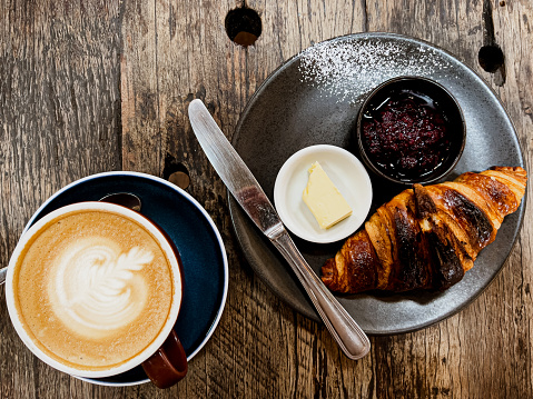 Horizontal high angle closeup photo of a freshly baked croissant with blueberry compote served on a round grey ceramic plate and a cup of latte coffee with foam art, on a rustic hand built wooden plank table in a cafe. Uralla near Armidale, New England high country, NSW in Autumn.