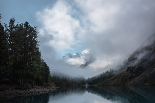 istock Tranquil scenery with snow castle in clouds. Mountain creek flows from forest hills into glacial lake. Snowy mountains in fog clearance. Small river and coniferous trees reflected in calm alpine lake. 1486178478