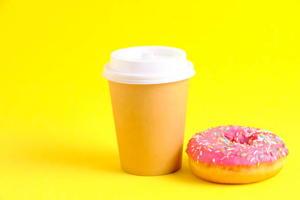 istock Disposable coffee cup and doughnut with pink icing yellow background. 1486183617