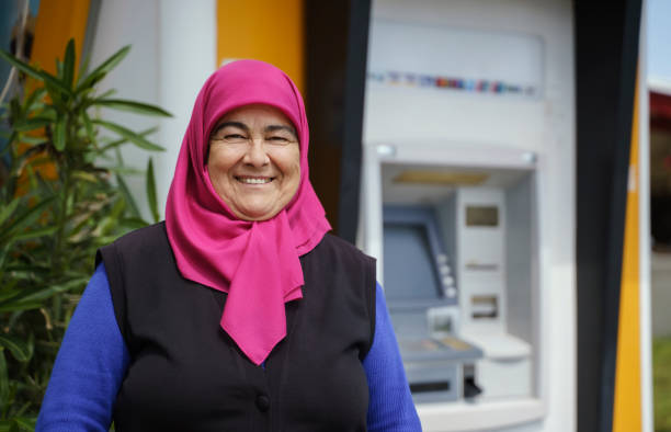Retired senior woman wearing a headscarf standing in front of a bank machine outdoors stock photo