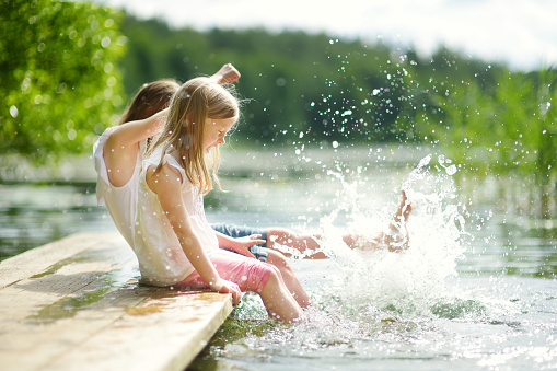 Two cute little girls sitting on a wooden platform by the river or lake dipping their feet in the water on warm summer day. Family activities in summer.